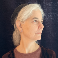A grey haired women looks to her left. She has a maroon shirt a black hat and a small silver earring.