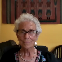 A white woman with white curly hair sitting in front of a yellow wall, maroon and teal ikat tapestry hangs behind her
